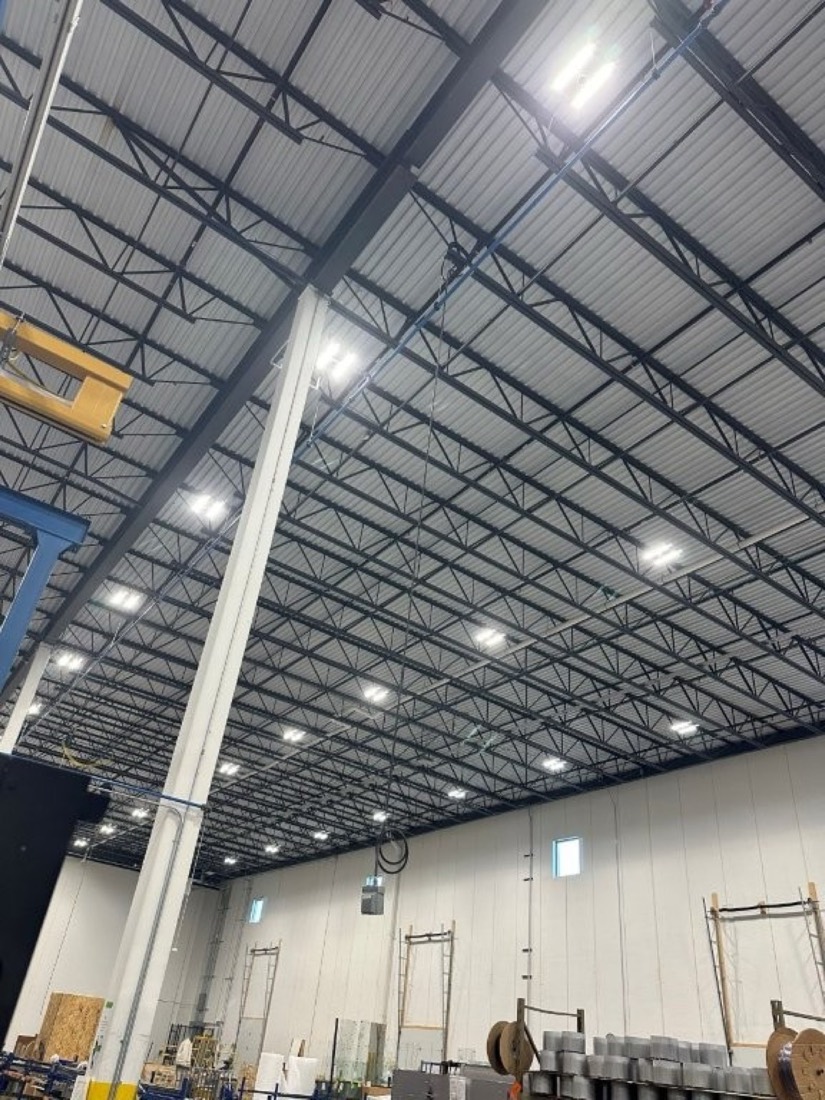 Rows of LED high-bay light fixtures on the open steel-joist ceiling of a warehouse.