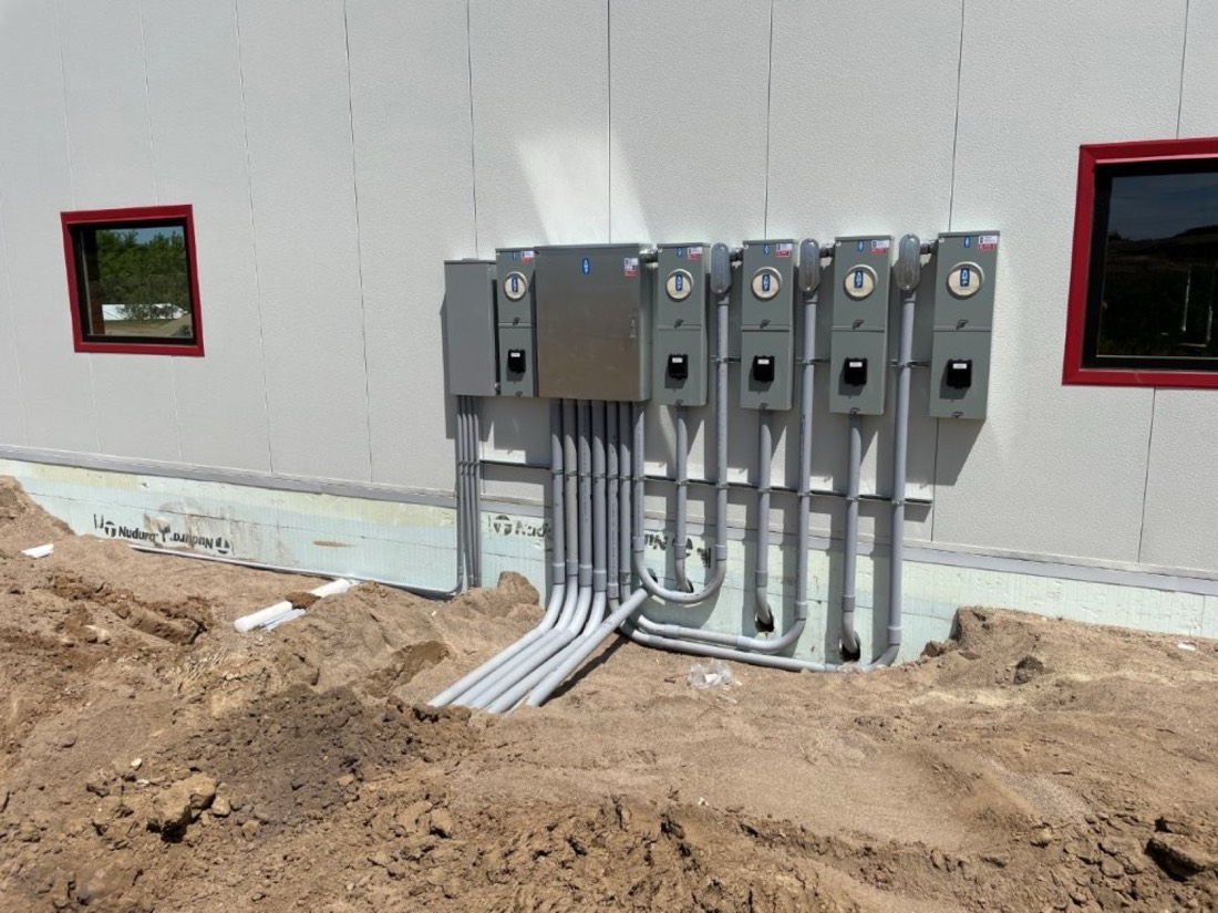 A bank of electrical meters and disconnects mounted on the exterior wall of a new commercial building.