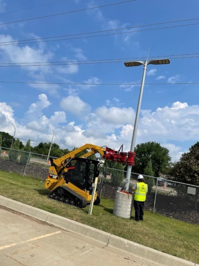 An electrician guiding a light pole onto its concrete base while a compact track loader with a hydraulic pole setter holds it upright.