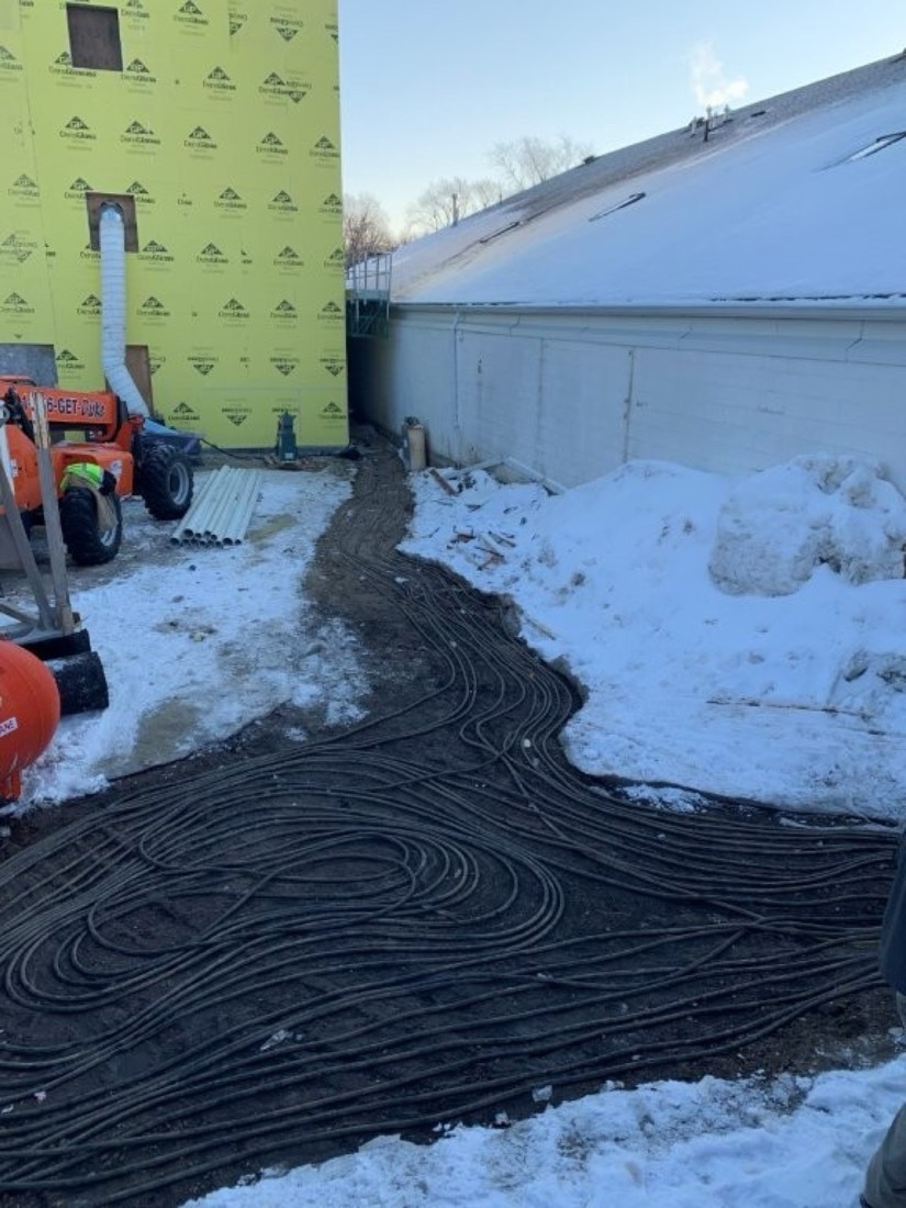 Heat loops laid in a serpentine pattern across frozen, snow-covered ground beside a commercial building under construction.